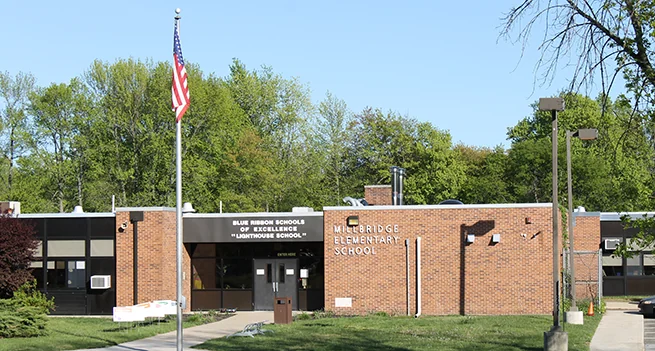 Single-story brick school building with American flag, entrance doors, and mature trees surrounding it