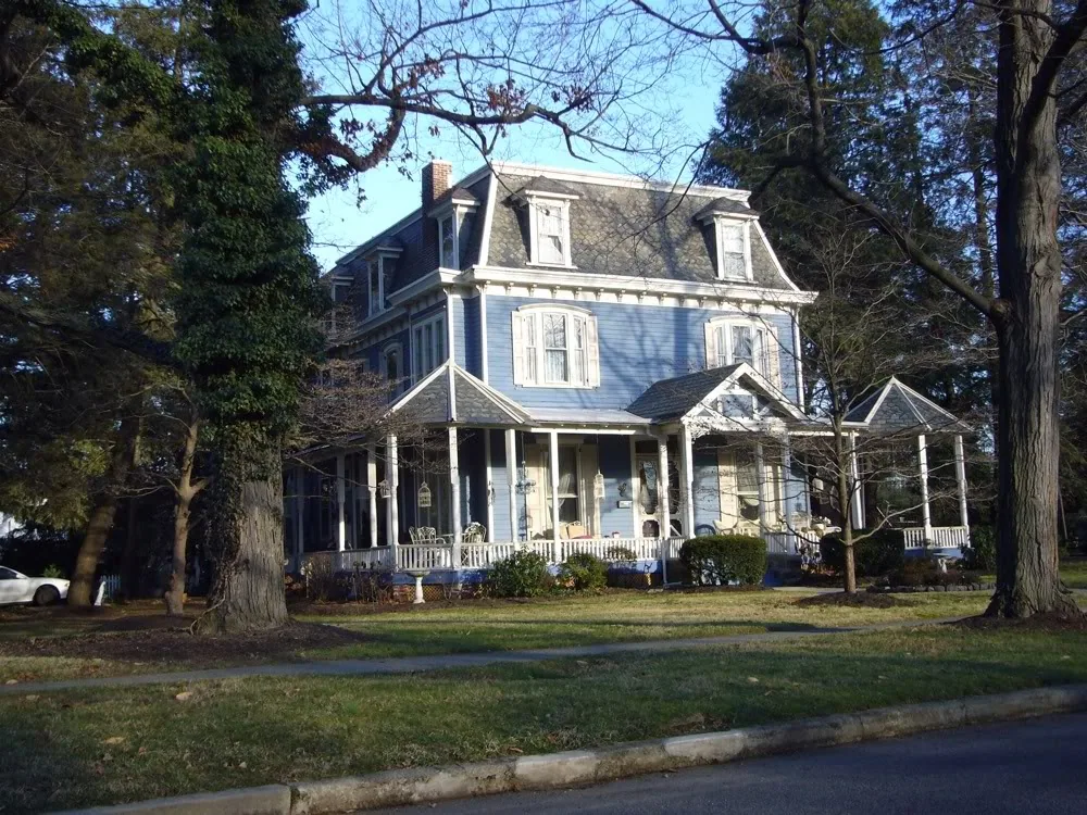 Victorian-style house with blue siding, mansard roof, front porch, and white trim surrounded by large trees