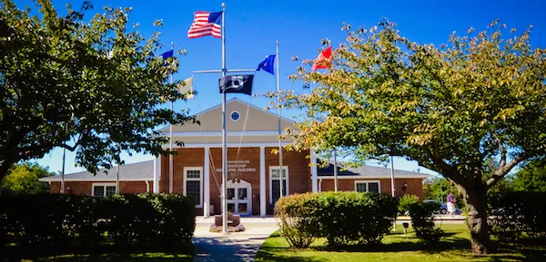Red brick town hall building with white columns and cupola, flying American flag and two other flags, surrounded by green landscaping and trees